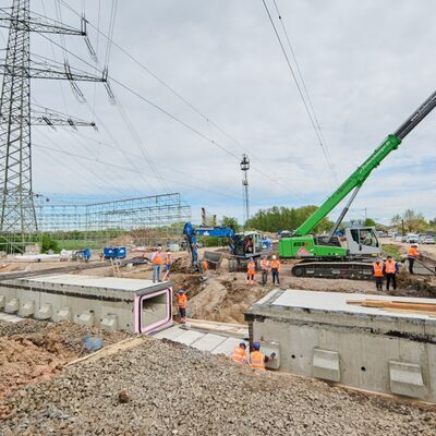 Zwei Betonblöcke für einen Durchlass unter den Gleisen liegen in einer Baugrube. Dahinter ein Bagger und ein Mobilkran sowie zahlreiche Bauarbeiter.